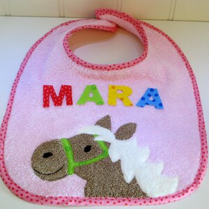 Könnte beinhalten: Rosa Baby-Lätzchen mit einem Pferde-Design und dem Namen "MARA" in bunten Buchstaben.