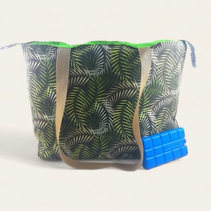 Könnte beinhalten: Eine Tragetasche mit tropischem Palmenblattmuster in Schwarz, Weiß und Grün. Die Tasche hat beige Träger und ein hellgrünes Innenfutter. Ein blauer Kühlakku liegt neben der Tasche.