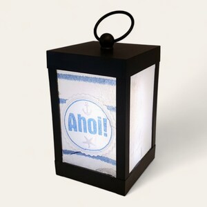 Könnte beinhalten: Eine schwarze Laterne mit einem weißen und blauen nautischen Design. Die Laterne hat einen Griff oben und steht "Ahoy!" auf der Vorderseite.