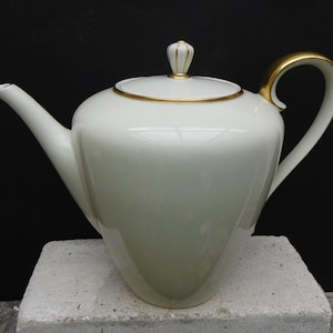 May include: A white porcelain coffee pot with a gold trim and a gold knob on the lid. The pot has a rounded body and a spout on the side.