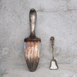 Puede incluir: Una antigua paleta para tartas de plata y una pequeña cuchara en forma de campana. La paleta para tartas tiene un mango largo y ornamentado y una hoja ancha y plana. La cuchara presenta un diseño de rosa en el mango. Ambos artículos se muestran sobre un fondo gris texturizado.