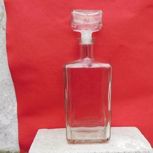 Puede incluir: Una decantador de vidrio transparente con cuerpo cuadrado y tapón cuadrado. La decantador está vacía y se encuentra sobre una superficie blanca.