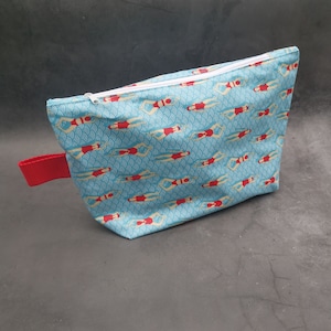 Könnte beinhalten: Eine blaue Reißverschlusstasche mit einem roten Griff. Die Tasche hat ein sich wiederholendes Muster von Schwimmern in roten und weißen Badeanzügen auf einem blau-weißen Wellenhintergrund.