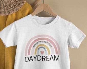 Puede incluir: Camiseta blanca con un diseño de arcoíris en colores pastel y el texto "darling I'm a nightmare dressed like a DAYDREAM". La camiseta está colgada en una percha de madera.