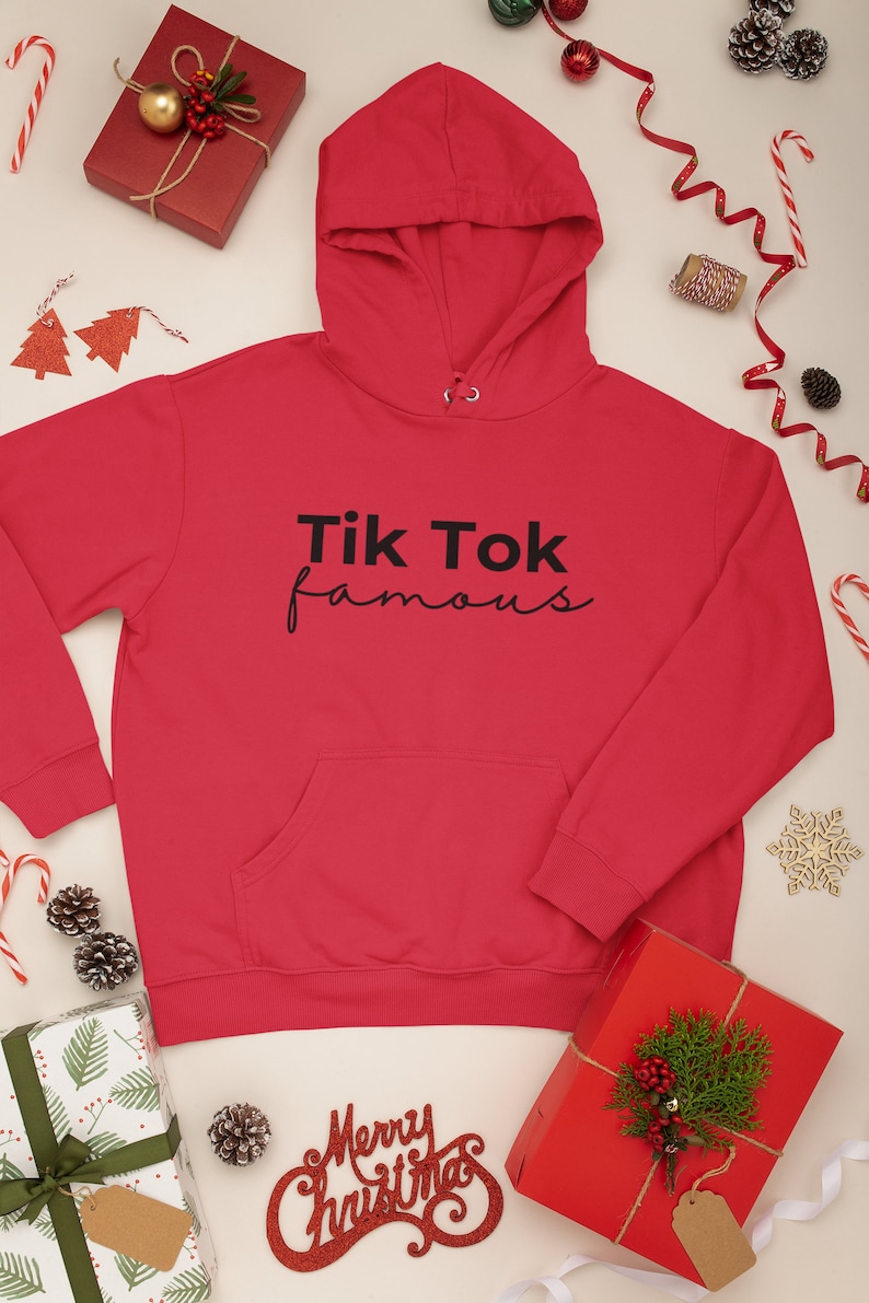 Puede incluir: Una sudadera con capucha roja con el texto "Tik Tok famous" en negro. La sudadera tiene un bolsillo delantero y una capucha con cord&oacute;n. La imagen est&aacute; rodeada de decoraciones navide&ntilde;as, incluyendo regalos envueltos, bastones de caramelo y pi&ntilde;as.