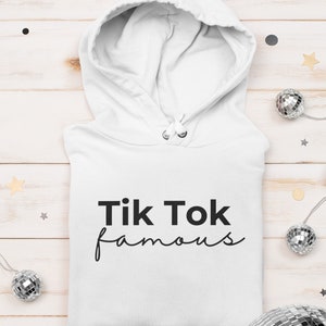 Puede incluir: Sudadera con capucha blanca con el texto negro "Tik Tok famous". La sudadera est&aacute; doblada sobre una superficie de madera clara, con elementos decorativos como bolas de discoteca y estrellas.