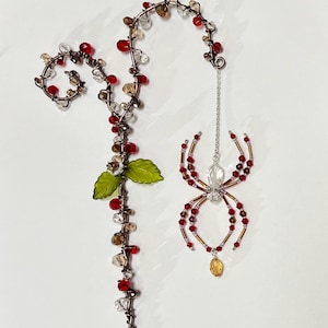 May include: A handcrafted beaded spider ornament with red, clear, and brown beads. The spider hangs from a silver chain. A beaded vine with red, clear, and brown beads and green leaves is also present.
