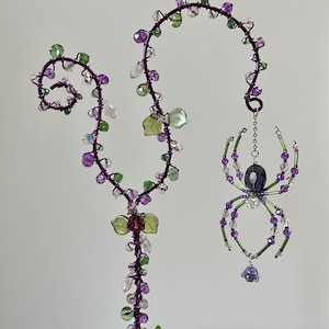 May include: A decorative wire sculpture featuring a purple vine with green and purple beads, and a hanging spider ornament. The spider has a purple body, green and purple legs, and a dangling purple bead. The sculpture is placed above a succulent plant.