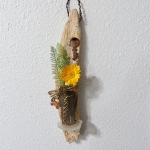 May include: A rustic wooden wall hanging with a small glass vase holding a single yellow flower. The vase is attached to the wood with twine and a rusty key is attached to the wood above the vase.
