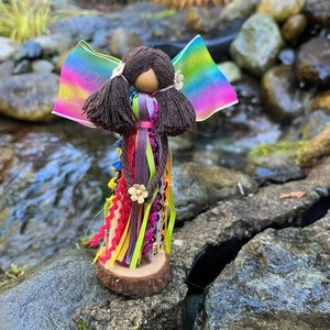May include: A colorful handmade doll with rainbow wings, brown yarn hair, and a wooden head. The doll is wearing a dress made of colorful ribbons and is standing on a wooden base.