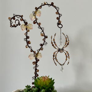 May include: A decorative metal spider with brown and clear beads hangs from a curved metal vine with leaf accents. The vine is adorned with brown and clear beads. A succulent plant is at the base.