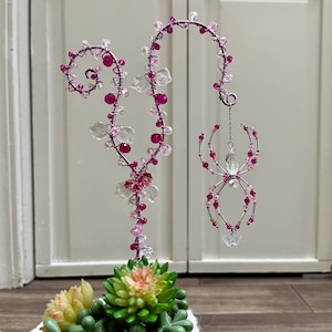 May include: A handcrafted wire art piece featuring a decorative spider and a floral design. The spider and floral elements are adorned with pink and clear beads. The base includes a small succulent arrangement.