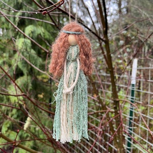 May include: A handmade doll ornament with a light green and white yarn dress, brown yarn hair, and a wooden head. The doll is hanging from a branch.