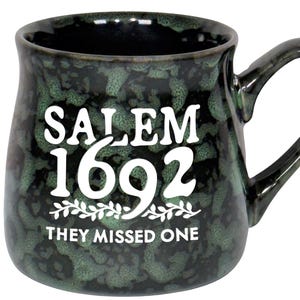 May include: A ceramic mug with a dark green speckled glaze. The mug features the text "SALEM 1692" in white, with "THEY MISSED ONE" below. The mug has a large handle and a wide opening.