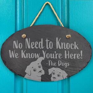 May include: A black oval slate sign with the text "No Need to Knock We Know You're Here! -The Dogs" and two dog silhouettes. The sign is hanging on a teal blue door.