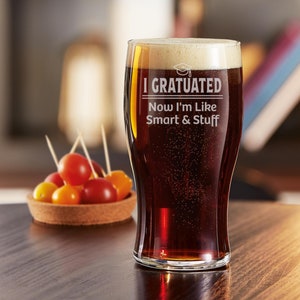 May include: A clear glass pint glass with a dark brown liquid inside. The glass has the text "I GRADUATED Now I'm Like Smart & Stuff" etched on the side.