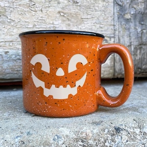 May include: Orange ceramic mug with a black rim and a white jack-o'-lantern face design. The mug has a speckled orange glaze.