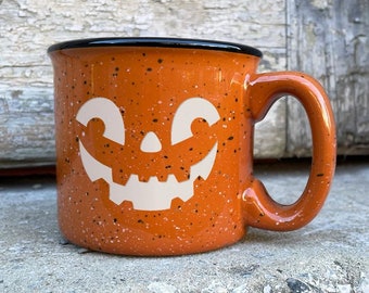 May include: Orange ceramic mug with a black rim and a white jack-o'-lantern face design. The mug has a speckled orange glaze.