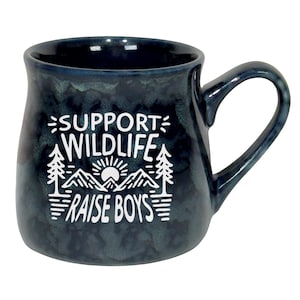 May include: A dark blue ceramic mug with a white graphic that reads "SUPPORT WILDLIFE RAISE BOYS". The design includes mountains, trees, and a sun. The mug has a curved handle and a speckled finish.