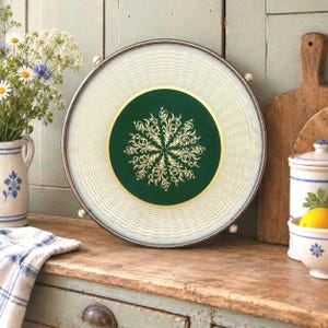 Könnte beinhalten: Ein rundes Serviertablett mit einer dunkelgrünen, kreisförmigen Mitte, die mit einem goldenen filigranen Blumenmuster verziert ist. Der äußere Rand besteht aus Milchglas mit goldenen Wellenlinien, eingefasst in einen Metallrahmen.