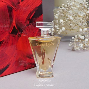 May include: A clear glass bottle of Champs-&Eacute;lys&eacute;es perfume with a gold label. The bottle is a miniature size and is sitting on a white surface. The text "Miniature Parfum Miniatur" is printed below the bottle.
