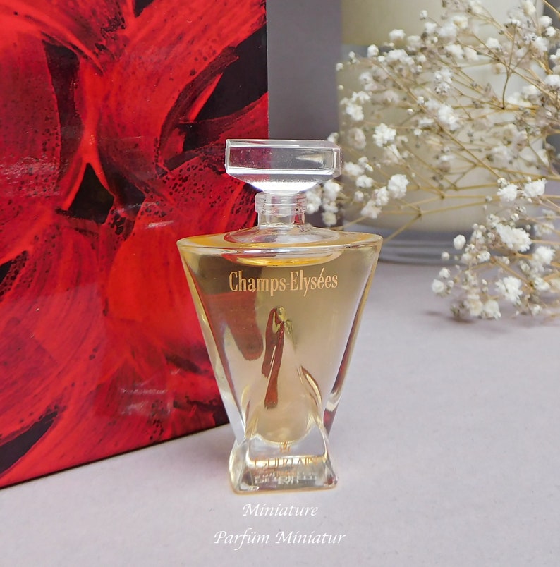May include: A clear glass bottle of Champs-&Eacute;lys&eacute;es perfume with a gold label. The bottle is a miniature size and is sitting on a white surface. The text "Miniature Parfum Miniatur" is printed below the bottle.