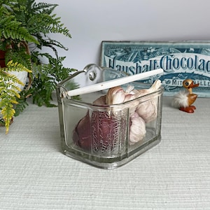 Pode incluir: Um recipiente de vidro transparente para alho com uma alça branca, contendo vários bulbos de alho. O recipiente tem formato triangular e um design decorativo na frente. Uma placa de chocolate vintage está ao fundo.