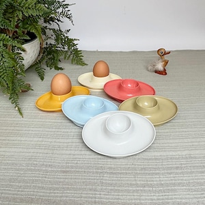 Peut inclure: Plusieurs coquetiers colorés, jaunes, roses, bleus et blancs, chacun contenant un œuf brun. Un petit canard en bois et une fougère en pot sont visibles en arrière-plan.