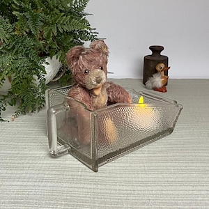 Peut inclure: Un ours en peluche rose vintage est assis dans un récipient en verre transparent texturé avec une poignée. Une bougie LED jaune est à l'intérieur du récipient. Une plante de fougère et d'autres objets décoratifs sont en arrière-plan.