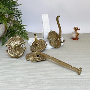 Op de afbeelding: Een verzameling antieke messing haken en een handdoekstang. De haken hebben sierlijke, decoratieve achterplaten en gebogen haken. De handdoekstang is rechthoekig met een decoratief uiteinde. Een klein houten eendenbeeldje staat op de achtergrond.
