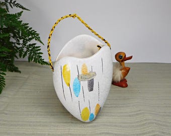 Op de afbeelding: Witte keramische wandpot met een gele touwgreep. De pot heeft een unieke vorm en is versierd met abstracte gele, blauwe en oranje vormen en zwarte lijnen. De pot heeft een label met de tekst "W. Keramik".