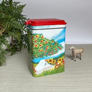 May include: A rectangular metal tin with a red lid and a colorful pastoral scene. The scene depicts an apple tree, sunflowers, and a figure in white. A small sheep figurine stands nearby.