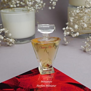 May include: A clear glass bottle of Champs-Elysees perfume by Guerlain. The bottle is a miniature size and has a gold liquid inside. The bottle is sitting on a red surface with the words "Miniature Parfum Miniatur" written on it.