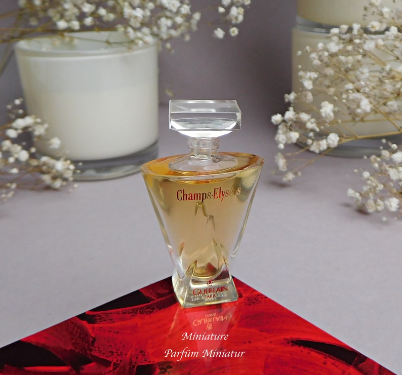 May include: A clear glass bottle of Champs-Elysees perfume by Guerlain. The bottle is a miniature size and has a gold liquid inside. The bottle is sitting on a red surface with the words "Miniature Parfum Miniatur" written on it.
