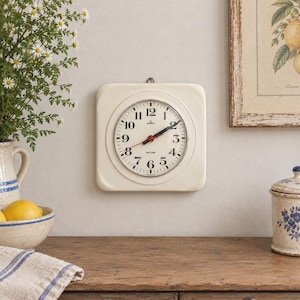Könnte beinhalten: Eine quadratische, cremefarbene Wanduhr mit rundem Zifferblatt, schwarzen arabischen Ziffern, silberfarbener Lünette sowie schwarzen Stunden- und Minutenzeigern und einem roten Sekundenzeiger. Auf dem Zifferblatt ist der Markenname Junghans zu sehen.