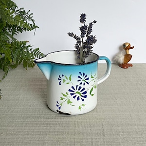 Könnte beinhalten: Eine Vintage-Kanne aus weißem Emaille mit blauem Ombre-Rand und Griff, verziert mit blauen und grünen Blumenmustern. Die Kanne enthält getrockneten Lavendel. Eine kleine Holz-Entenfigur befindet sich im Hintergrund.