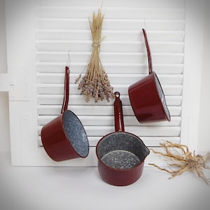 Può includere: Tre pentole di smalto rosso con interni grigi macchiati appese a una parete bianca a doghe. Le pentole hanno manici lunghi e sono di dimensioni diverse.