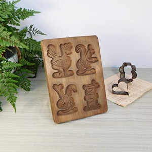 Könnte beinhalten: Eine antike Holz-Keksschablone mit vier verschiedenen Designs: ein Hahn, ein Eichhörnchen, ein Kaninchen und eine Garnrolle. Die Schablone ist aus dunkelbraunem Holz und hat eine rechteckige Form.