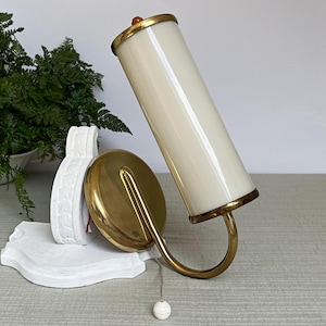 Può includere: Una lampada da parete con paralume cilindrico in vetro bianco sporco sostenuto da una struttura in ottone. L'apparecchio è fissato a una base decorativa bianca e include una piastra circolare in ottone con cordino e un piccolo interruttore.