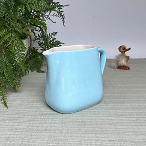 Op de afbeelding: Een lichtblauwe keramische kan met een witte binnenkant. De kan heeft een gebogen lichaam en een handvat. Op de achtergrond staat een klein houten eendenbeeldje met een pluizige witte borst.