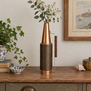 Puede incluir: Un jarrón cilíndrico alto con una sección superior de cobre pulido liso y una base de metal oscuro texturizado con un patrón martillado vertical. Un asa de metal dorado fino con un agarre de madera oscura está unida al costado del jarrón.