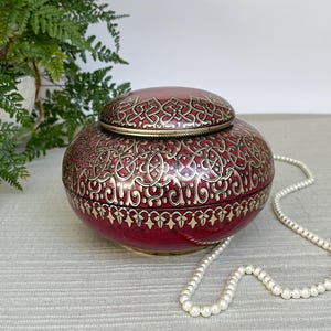 Op de afbeelding: Een ronde, rode decoratieve doos met een deksel, met gouden filigraanpatronen. De doos staat naast een witte parelketting. Een groene plant staat op de achtergrond.