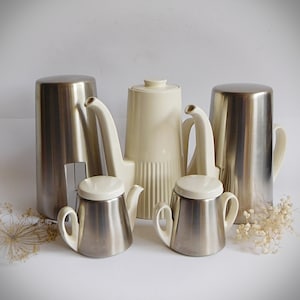 Op de afbeelding: Een set van vijf zilveren en witte keramische koffie- en theepotten. De set bevat een grote koffiepot, een kleinere theepot, een roomkan, een suikerpot en een bijpassende roomkan.