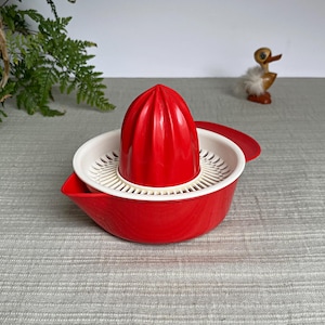 Op de afbeelding: Rode en witte citruspers met een conische ruimer en een zeef. De pers is gemaakt van plastic en heeft een klassiek ontwerp. De pers is ontworpen om sap uit citrusvruchten te halen. Een decoratief eendfiguur staat op de achtergrond.