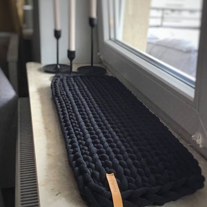 Könnte beinhalten: Schwarzes gestricktes Fensterbankkissen mit einem braunen Lederanhänger mit der Aufschrift "Sagittaria".