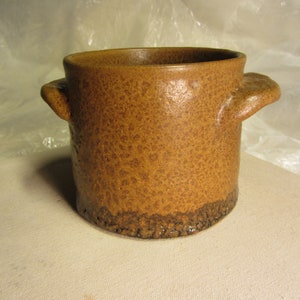 Pode incluir: Um recipiente de cerâmica artesanal com uma superfície texturizada e salpicada em tons de castanho e bege. Apresenta duas pequenas pegas arredondadas e uma base de textura rugosa castanho escuro. Esta peça de cerâmica é adequada para vários usos.