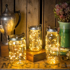 May include: Three clear glass mason jars with silver lids and handles, filled with warm white fairy lights. The jars are arranged on a wooden surface, with a silver teapot and a green vase with flowers in the background. The jars are lit up.