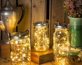 Könnte beinhalten: Drei klare Einmachgläser mit silbernen Deckeln und Griffen, gefüllt mit warmweißen Lichterketten. Die Gläser sind auf einer Holzoberfläche angeordnet, mit einer silbernen Teekanne und einer grünen Vase mit Blumen im Hintergrund. Die Gläser sind beleuchtet.