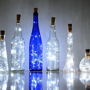 May include: Six glass bottles with cork tops, each containing a string of white LED lights. The bottles are arranged in a row on a white surface. The bottles are clear glass, except for one which is blue.
