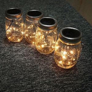 May include: Four glass jars with solar-powered lights inside. The jars have metal lids with a solar panel on top. The lights are on and casting a warm glow.